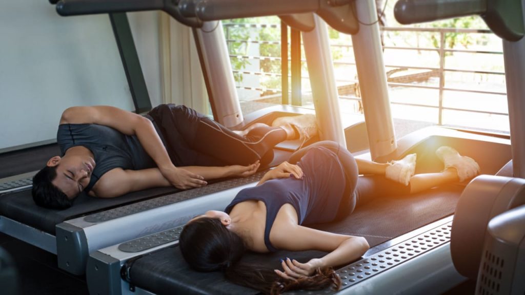 A man and a woman are lying down on treadmills, taking a break.