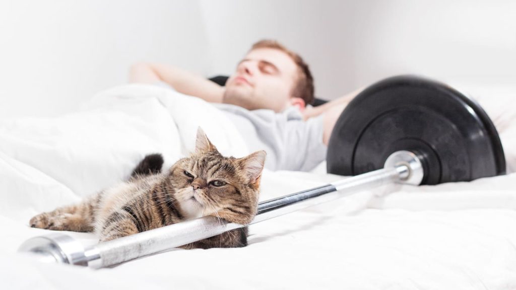 The athlete shows the importance of sleep after tough exercise while his cat is enjoying a lazy day.